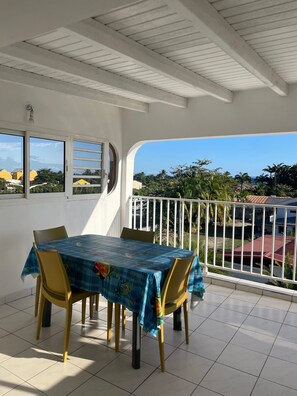 A covered patio with a table and chairs, a view of the sea, and a white railing. - RESIDENCE LE LAGON (Sainte-Anne)