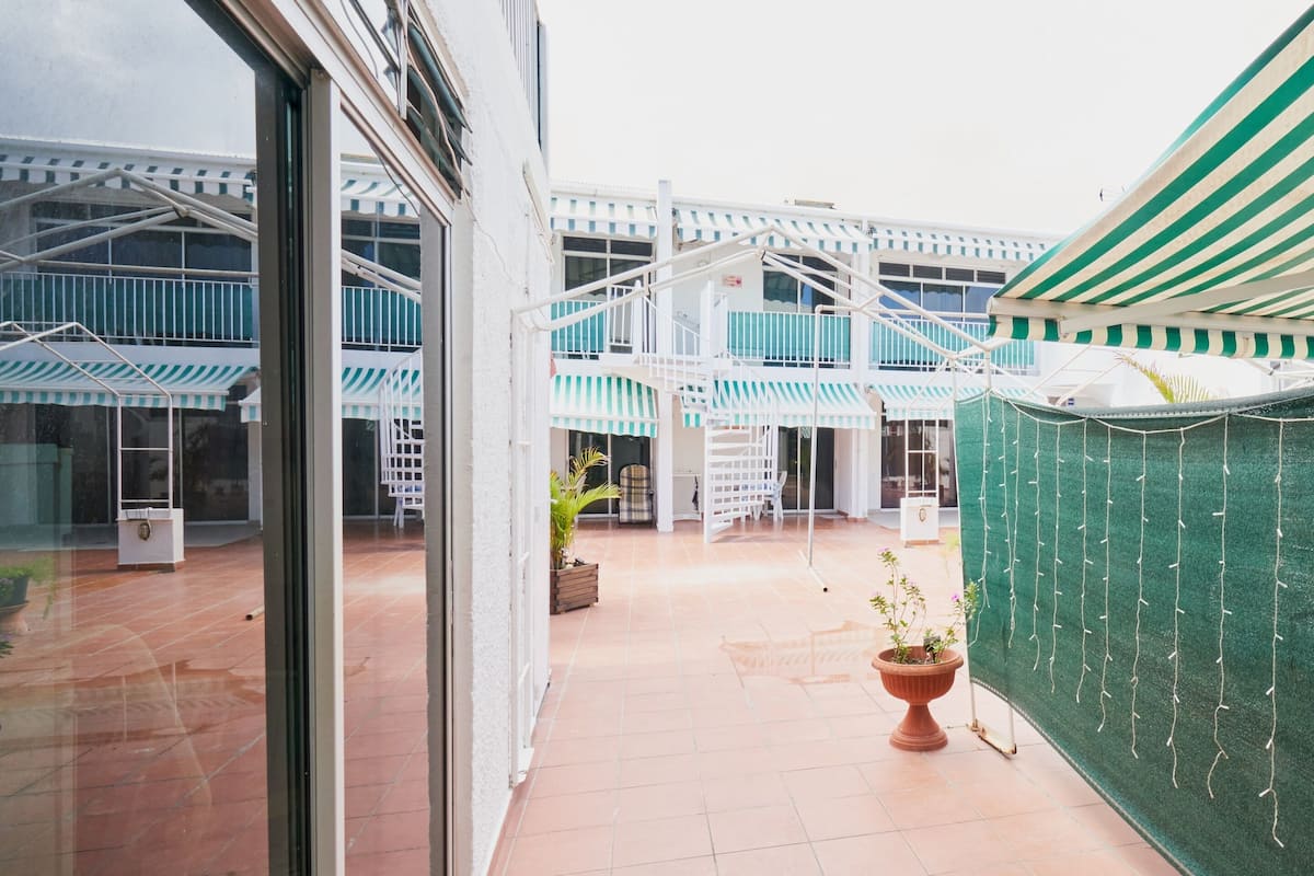 A spacious outdoor area with a tiled floor, a green plant, and a white building with multiple balconies.