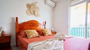 Quarto com cama de madeira, duas mesas de cabeceira, ar condicionado fixado na parede e janela com vista.