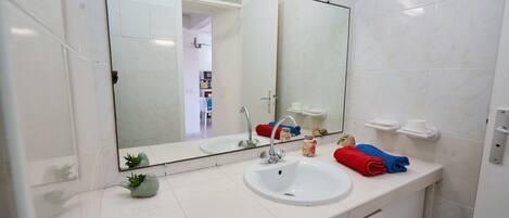 Um banheiro com pia branca, um espelho grande, porta-toalhas com toalhas vermelhas e azuis, e uma pequena planta no balcão.