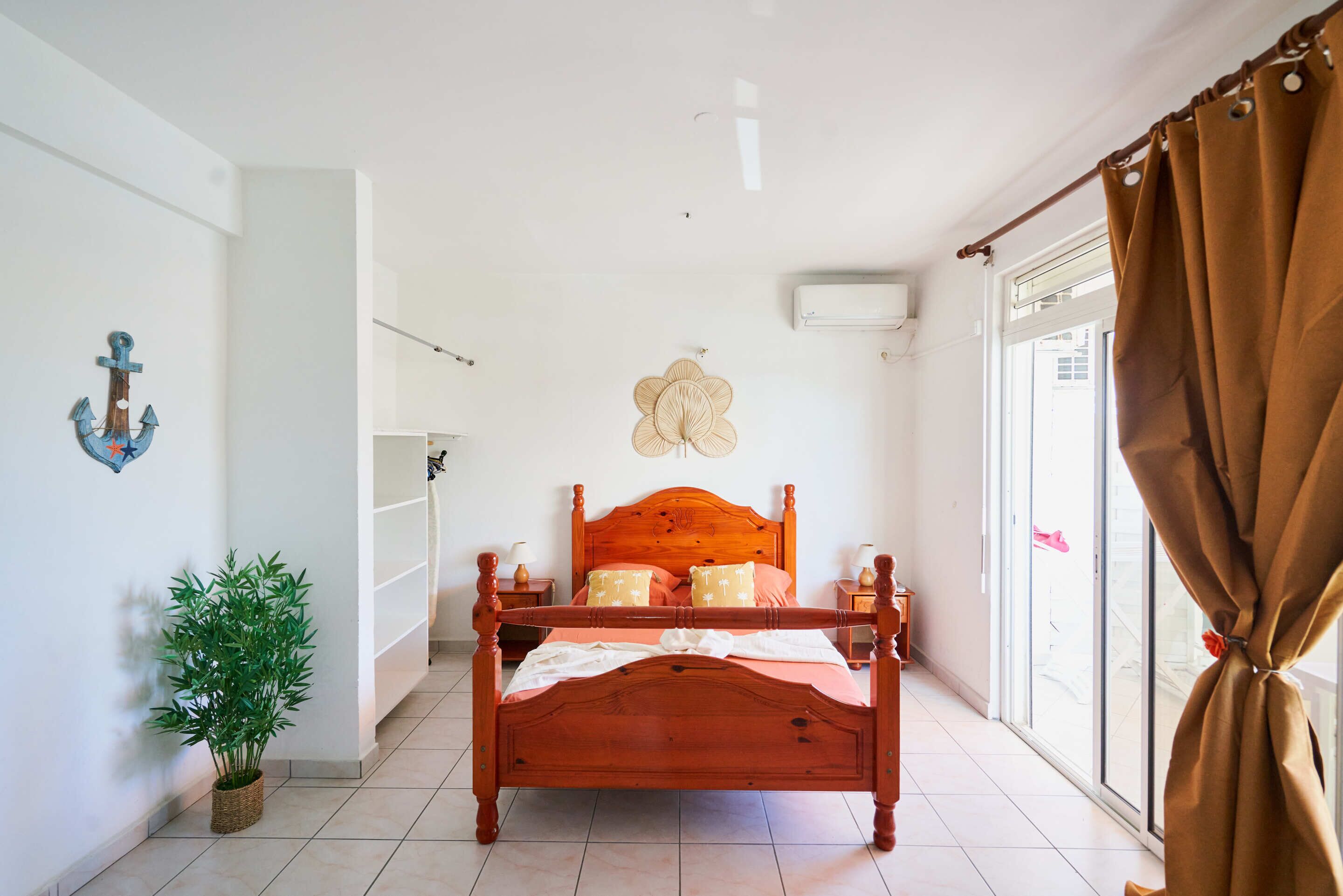 Une chambre à coucher avec un lit en bois, une plante en pot, un climatiseur fixé au mur et une fenêtre avec des rideaux.