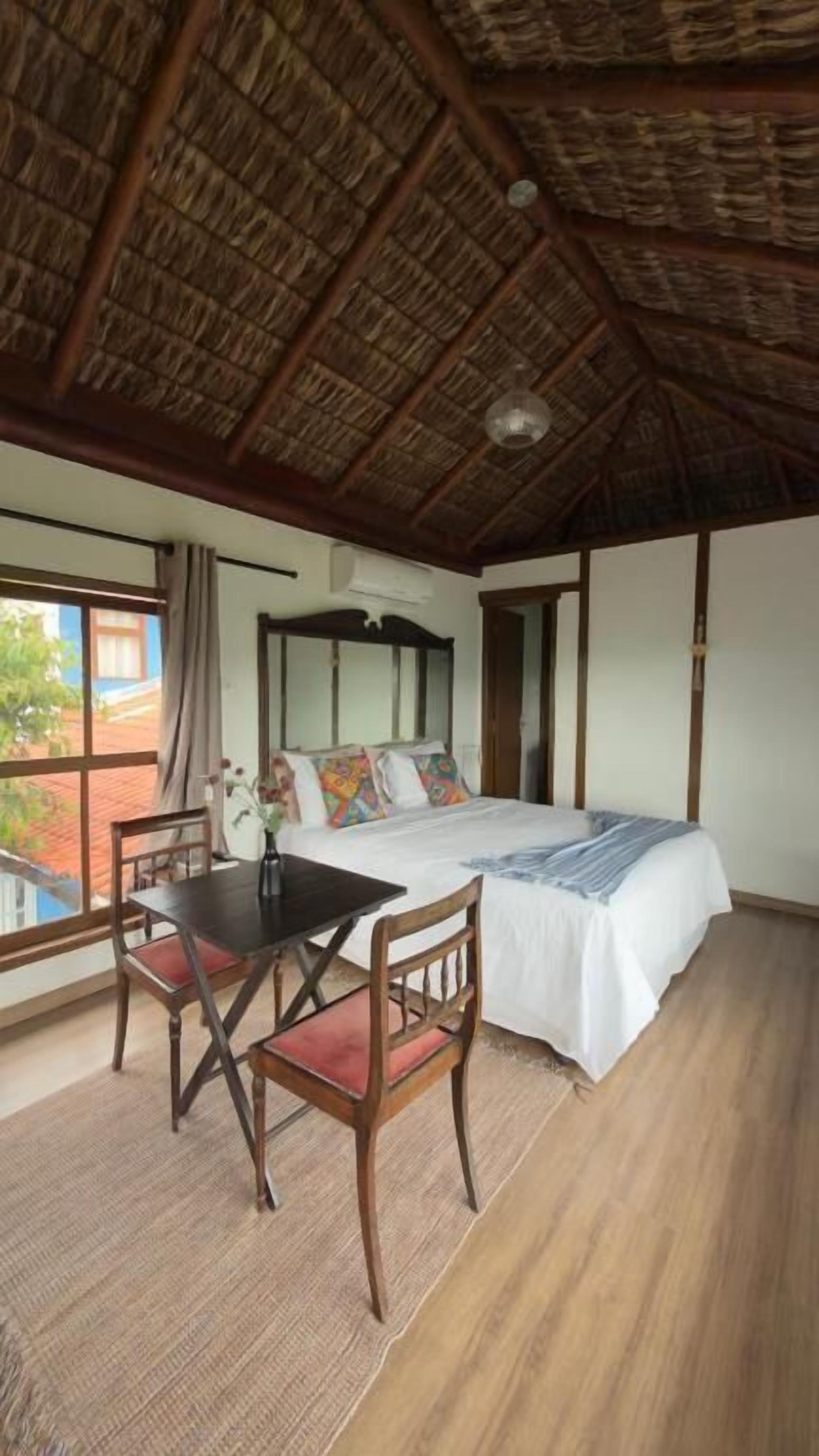 A room with a bed, a table, chairs, a window, and a thatched roof.