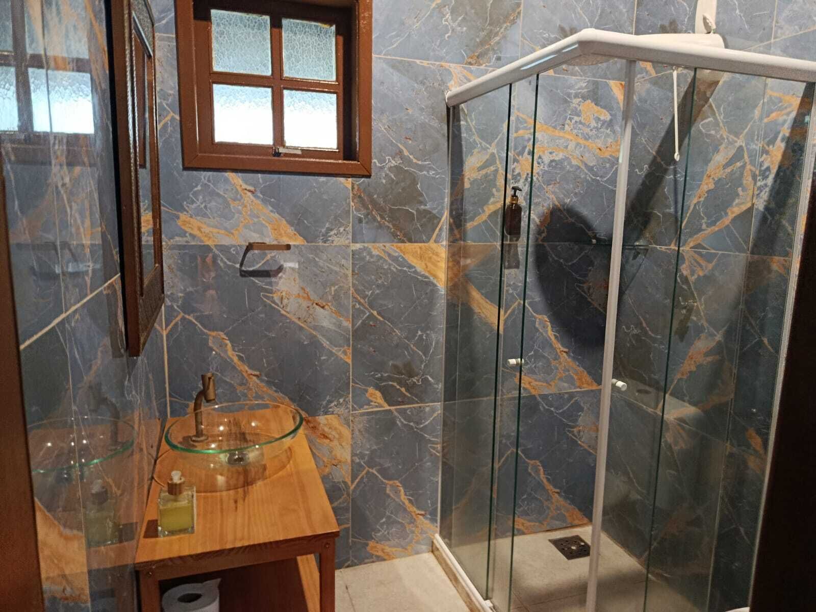 Een badkamer met een douche met glazen wand, een houten wastafelmeubel met een wasbak van glas en een raam boven de wasbak.