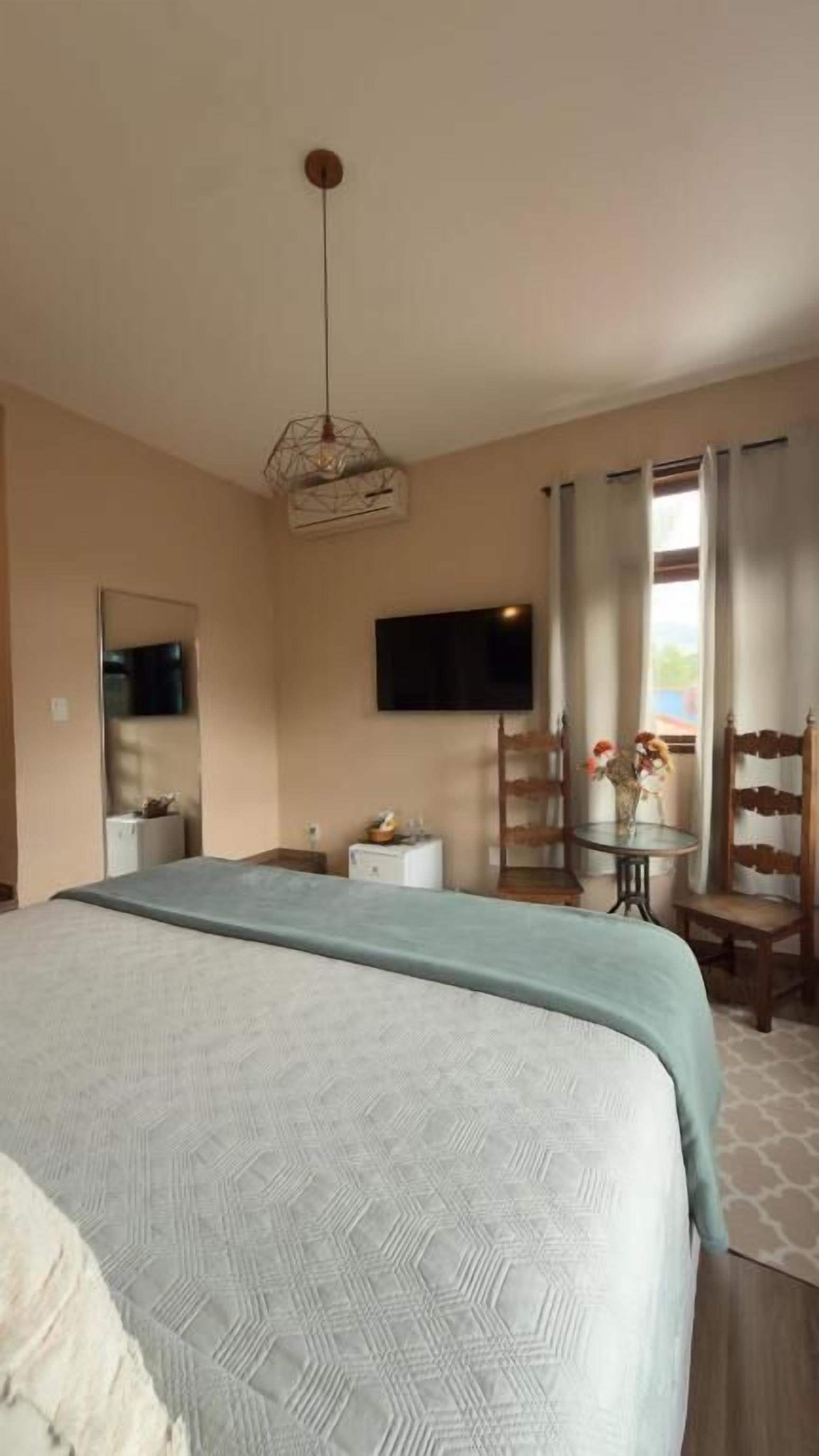 A bedroom with a bed, a TV mounted on the wall, a desk with a chair, and a window with curtains.