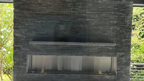 En eldstad i sten med en glasfront, placerad i ett modernt utomhusområde med en takkonstruktion i metall.