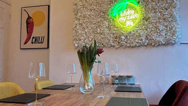 Ein Essbereich mit einem Holztisch, einer Neonreklame, einem gerahmten Chili-Gemälde und einer Vase mit Blumen.