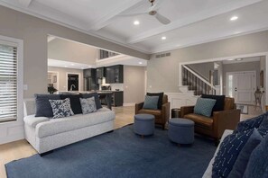 Uma sala de estar espaçosa com um sofá cinzento, duas poltronas de couro castanho e pufes. É visível uma cozinha ao fundo.