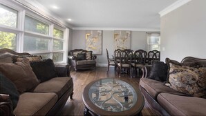A living room with a brown sofa, a glass coffee table, and a dining area with wooden chairs and a table. - Great Home_Sleeps10_ Large Yard_Room for Everyone (Overland Park)