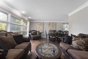 A living room with a brown sofa, a glass coffee table, and a dining area with wooden chairs and a table. - 3Queens 2Kings, w/Large Backyard,World Cup Ready (Overland Park)