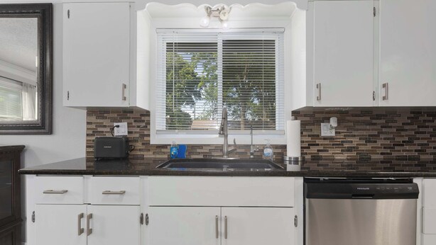 A kitchen with white cabinets, a stainless steel dishwasher, a window with blinds, and a backsplash with brown tiles. - Great Home_Sleeps10_ Large Yard_Room for Everyone (Overland Park)