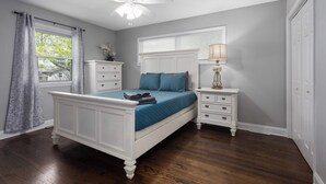 A bedroom with a bed, a nightstand, a lamp, a ceiling fan, a window with curtains, and a door. - Great Home_Sleeps10_ Large Yard_Room for Everyone (Overland Park)