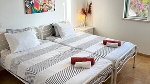 Ein Doppelbett mit gestreifter Bettwäsche, ein farbenfrohes Vogelgemälde an der Wand, eine Nachttischlampe und ein Fenster mit Blick.