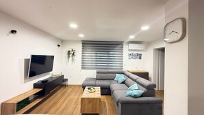 A modern living room with a flat-screen TV, grey sofa, wooden coffee table, and a wall-mounted clock.