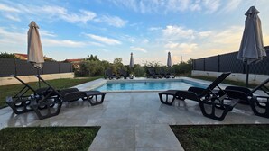 A swimming pool with lounge chairs and umbrellas, surrounded by a paved patio and a grassy area.