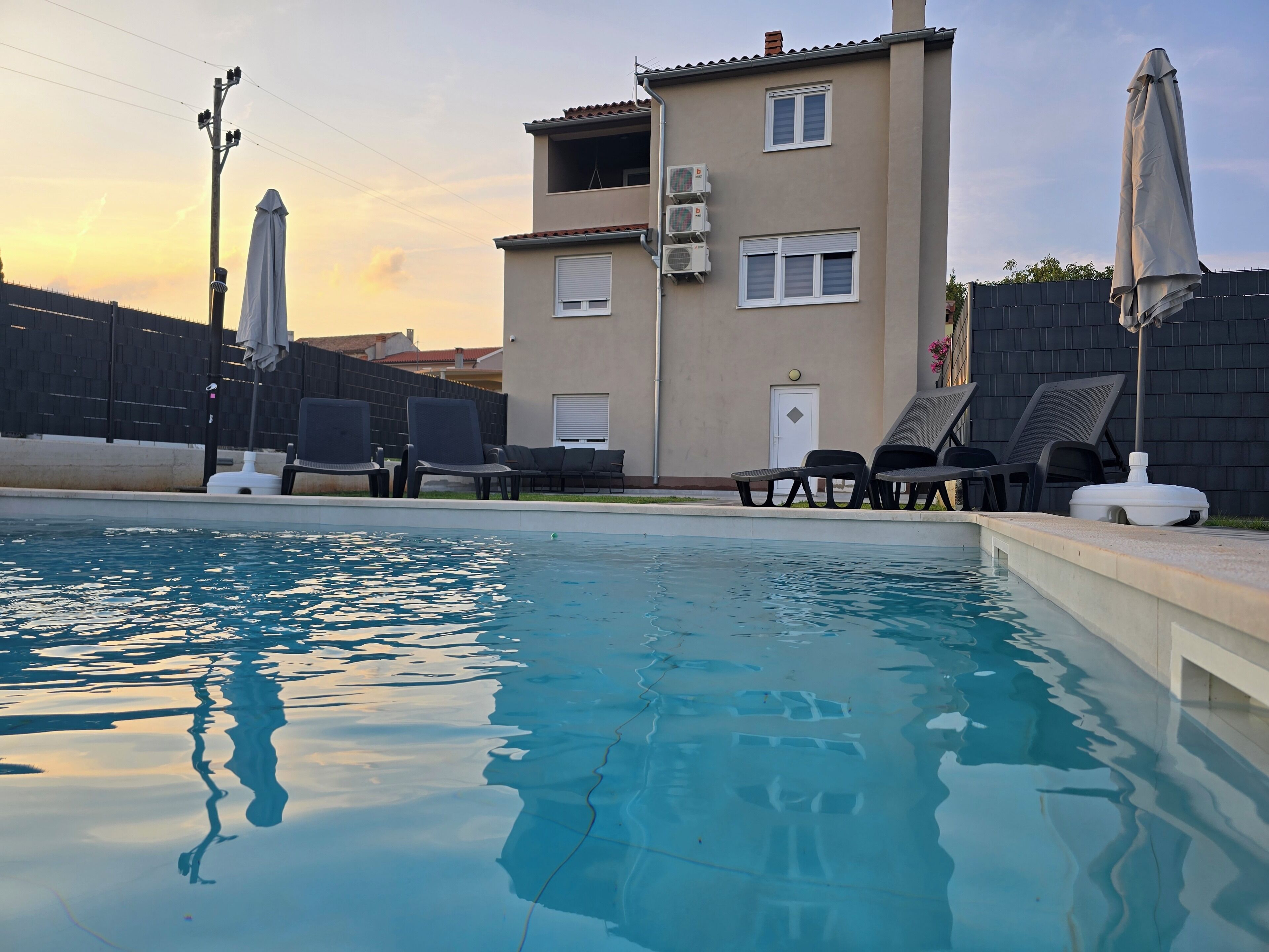 A swimming pool with lounge chairs and umbrellas, a building with air conditioning units, and a fence.