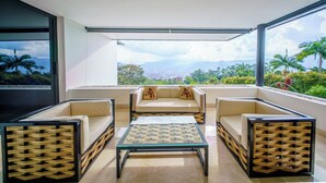 Un balcón moderno con mesa de vidrio y asientos acolchados, que ofrece una vista panorámica de una ciudad y montañas.