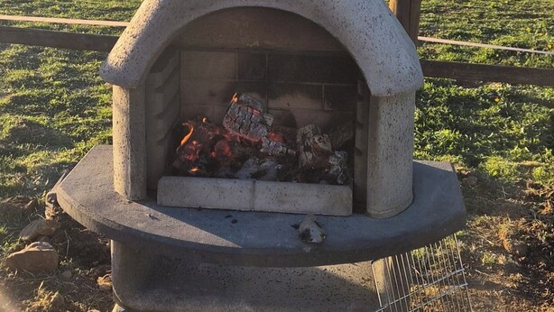 Ein steinerner Pizzaofen im Freien mit Feuer, umgeben von einer Wiese und einem Holzzaun.