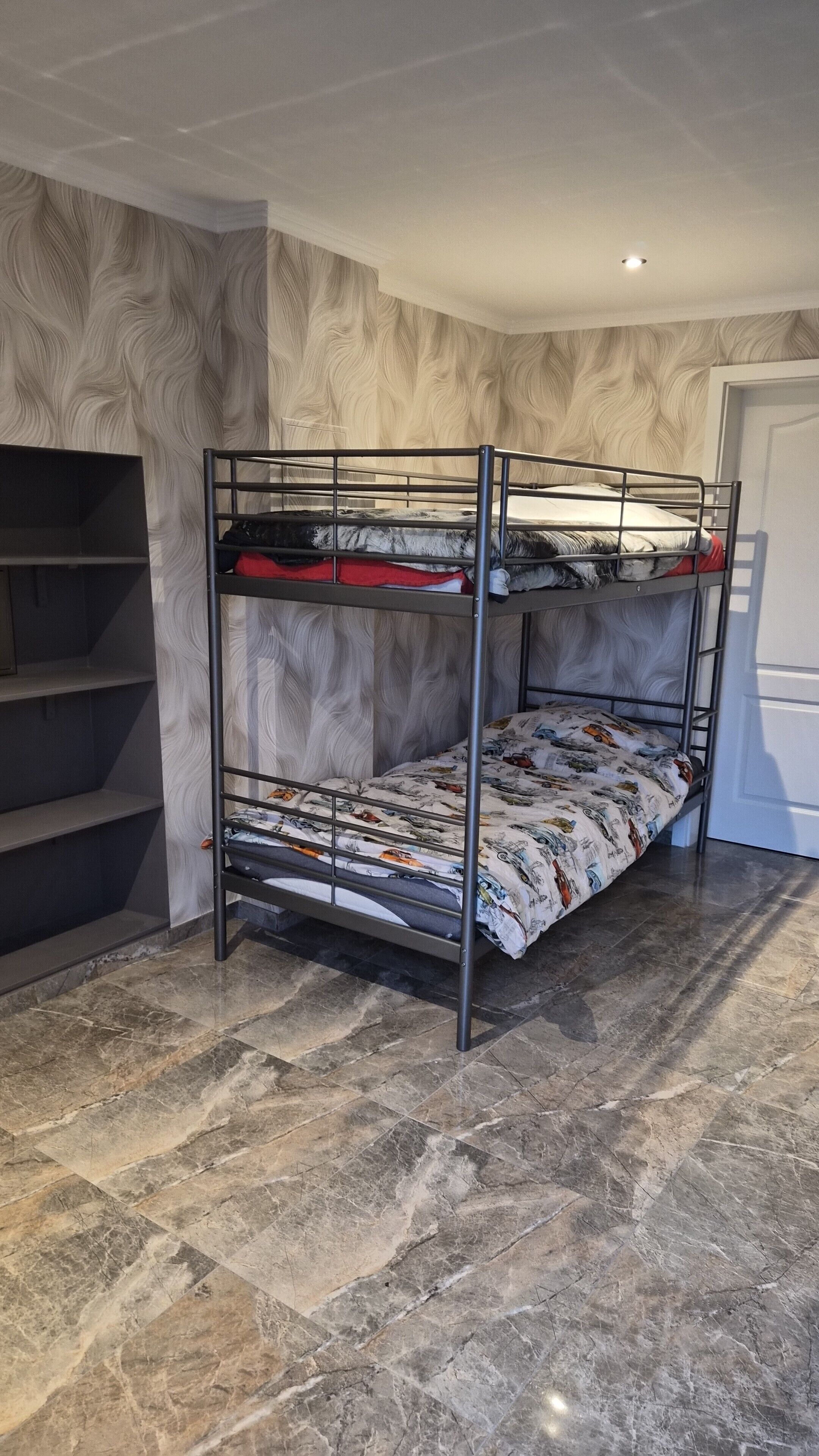 Un lit superposé avec une couvre-lit à motifs, une étagère et un sol carrelé en marbre, dans une pièce aux murs texturés.