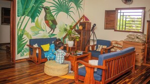 Una habitación con piso de madera, un sofá, una mesa de centro, una silla, una mesita con una planta y una ventana con vista a la vegetación.