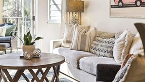 Ein Wohnzimmer mit rundem Holztisch, einer Couch mit vielen Kissen, einer Zimmerpflanze und einer Lampe.