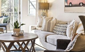 Ein Wohnzimmer mit rundem Holztisch, einer Couch mit vielen Kissen, einer Zimmerpflanze und einer Lampe.