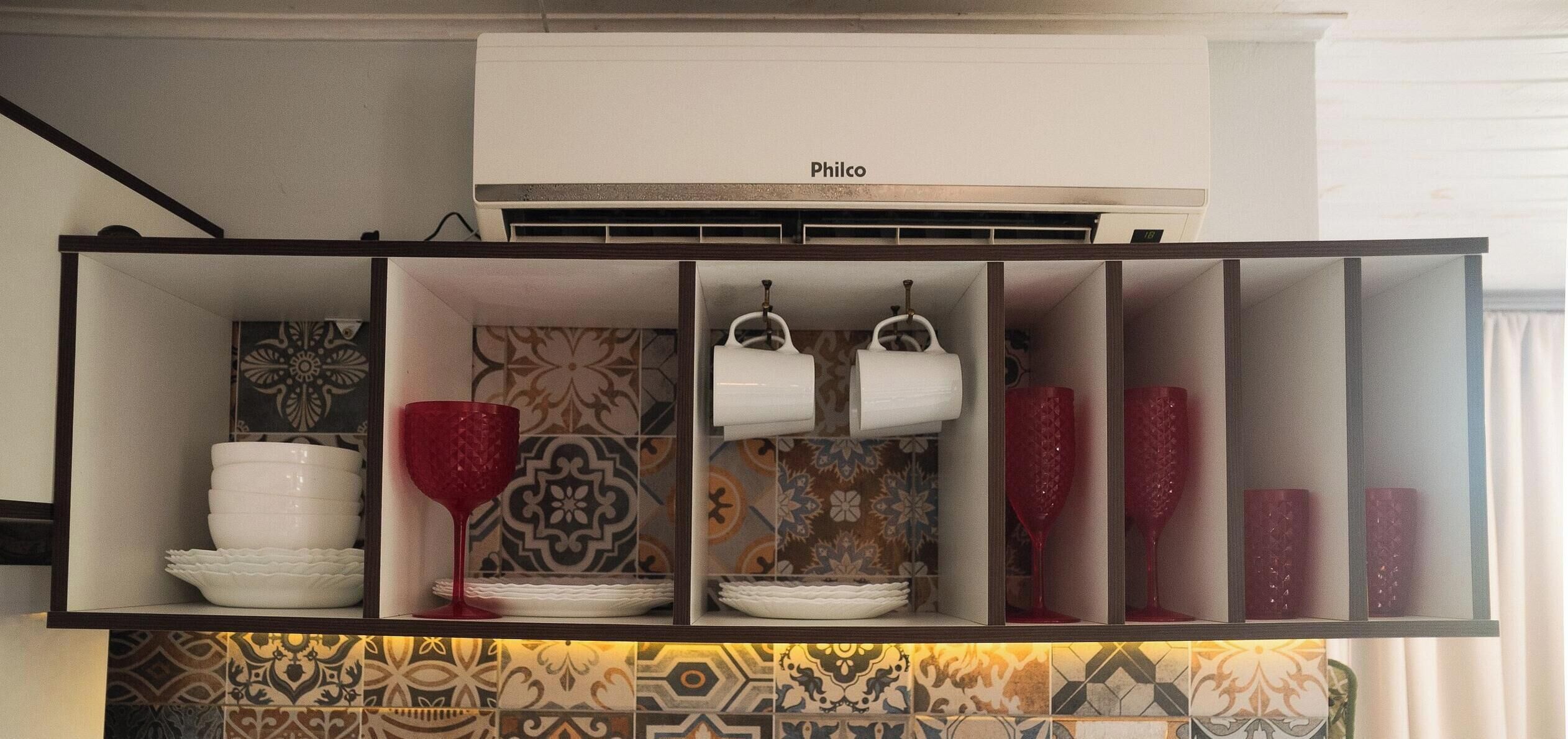 Cozinha com ar condicionado, azulejos estampados e prateleiras com louças brancas e copos de vinho tinto.