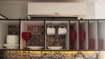 Cozinha com ar condicionado, azulejos estampados e prateleiras com louças brancas e copos de vinho tinto.