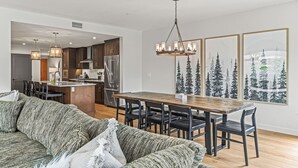 A modern kitchen with a central island, dining area with a wooden table and chairs, and a living room with a green sofa. - 1650 sq ft 2BR Corner Condo 303 (Canmore)