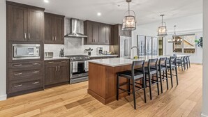 A modern kitchen with dark wood cabinets, stainless steel appliances, and a central island with bar stools. - 1650 sq ft 2BR Corner Condo 303 (Canmore)