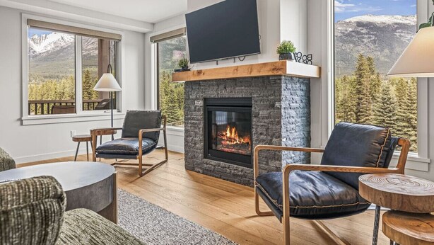 A modern living room with a fireplace, comfortable seating, a flat-screen TV, and large windows with mountain views. - 1650 sq ft 2BR Corner Condo 303 (Canmore)