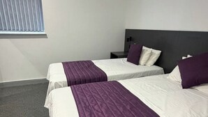 A hotel room with two beds, a headboard, a nightstand, and a window with blinds. - Commercial Hotel - Standard Twin Room Shared (Middlesbrough)