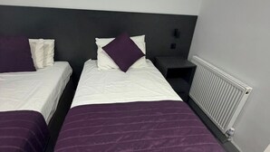 A hotel room with two beds, each with white bedding and purple accents, a grey headboard, a bedside table, and a radiator. - Commercial Hotel - Standard Twin Room Shared (Middlesbrough)