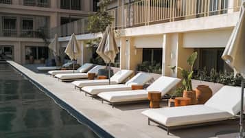 Área da piscina com espreguiçadeiras e guarda-sóis, piscina cristalina e um edifício com varandas ao fundo.
