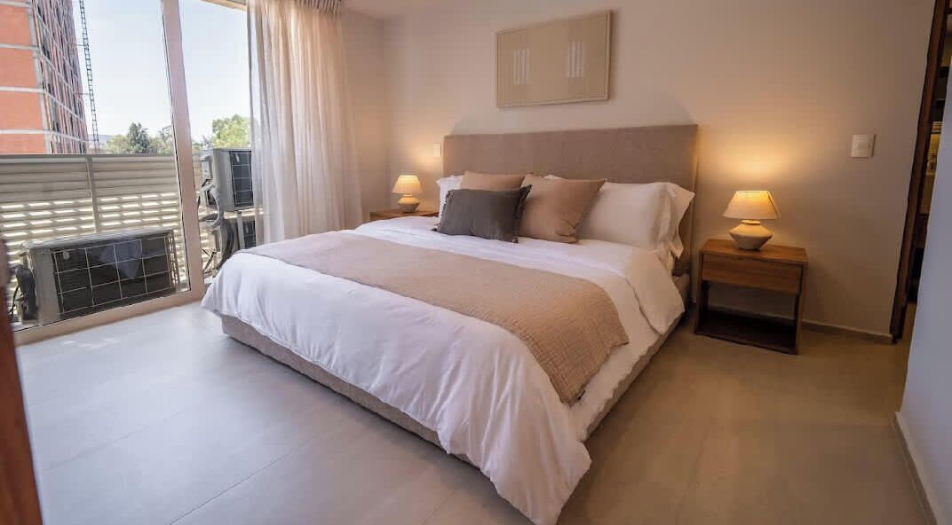 A hotel room with a large bed, two bedside tables with lamps, a view of the outside, and a window with curtains.