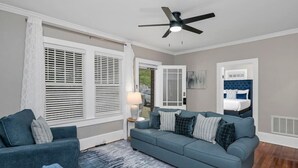Ein Zimmer mit einem blauen Sofa, einem Sessel, einem Bett mit blauer Kopfteil, einem Deckenventilator und einem Fenster mit Jalousien.