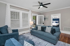 Una habitación con un sofá azul, una silla, una cama con cabecera azul, un ventilador de techo y una ventana con persianas.