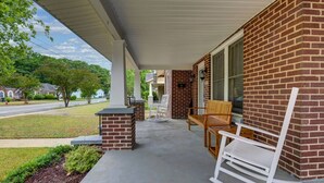 Eine überdachte Veranda mit Backsteinwand, Betonboden und Gartenmöbeln, darunter ein Schaukelstuhl und eine Bank.