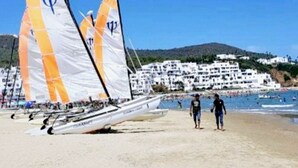 Un voilier aux voiles orange sur une plage de sable, une personne qui marche et, au loin, des bâtiments blancs.
