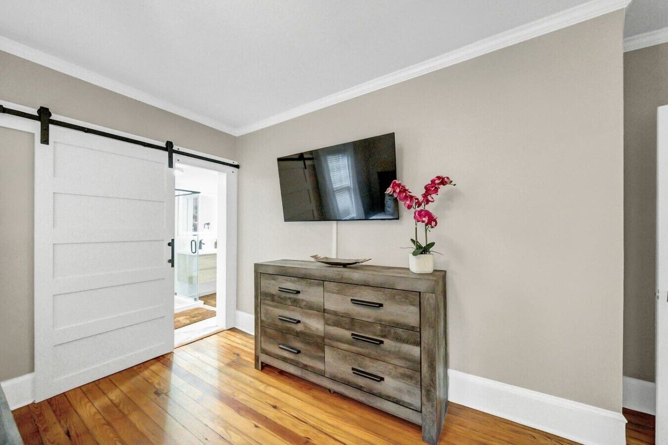 A room with a wooden floor, a sliding door, a grey dresser, a flat-screen TV, and a potted plant.