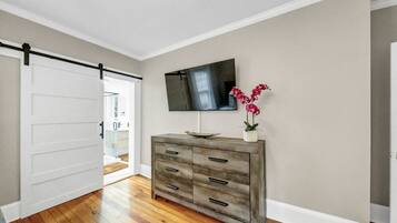 A room with a wooden floor, a sliding door, a grey dresser, a flat-screen TV, and a potted plant.