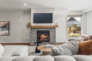 A living room with a fireplace, a flat-screen TV, and a view of a building through a window. - 1550 sq ft 2BR Premier Condo 302TML (Canmore)