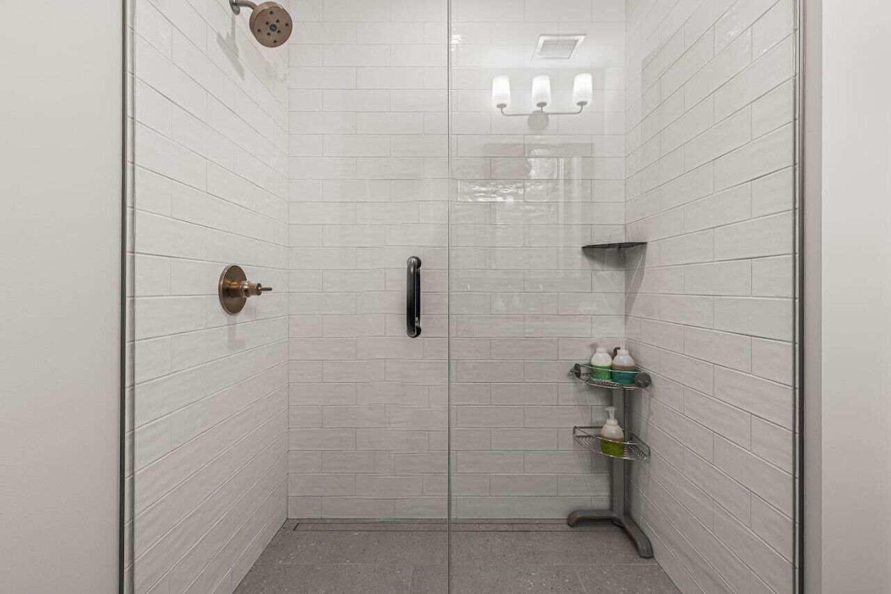 A walk-in shower with white tiled walls, a glass door, a showerhead, a towel rack, and a shelf with toiletries.