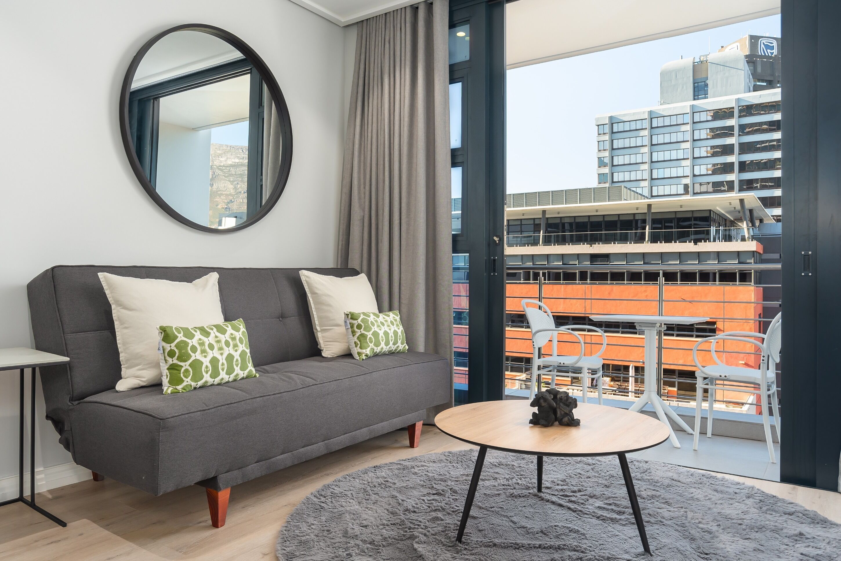 A modern living room with a grey sofa, a round coffee table, and a view of a cityscape through large windows.
