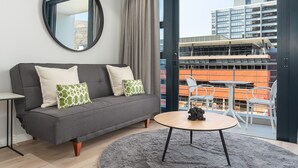 Ein modernes Wohnzimmer mit einem grauen Sofa, einem runden Couchtisch und Blick auf eine Stadtlandschaft durch große Fenster.