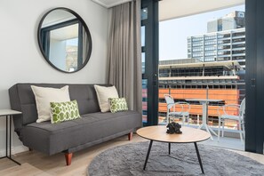 Ein modernes Wohnzimmer mit einem grauen Sofa, einem runden Couchtisch und Blick auf eine Stadtlandschaft durch große Fenster.