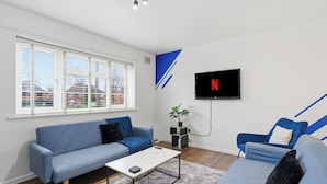 A modern living room with a sofa, a coffee table, and a TV mounted on the wall. - Contractor/Family Friendly 4 Bedroom House w/WIFI & Free Parking (West Bromwich)