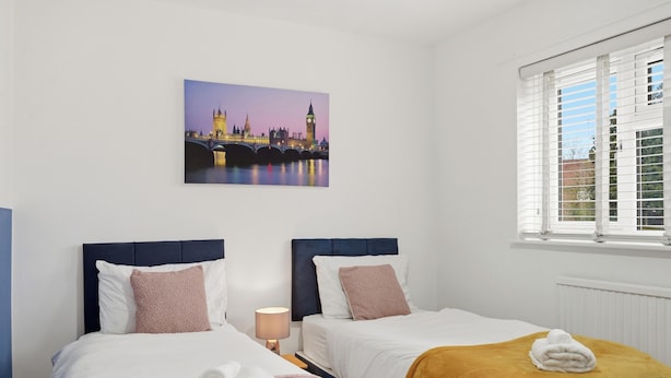 A hotel room with two beds, a window with blinds, a lamp, and a cityscape painting on the wall. - Contractor/Family Friendly 4 Bedroom House w/WIFI & Free Parking (West Bromwich)