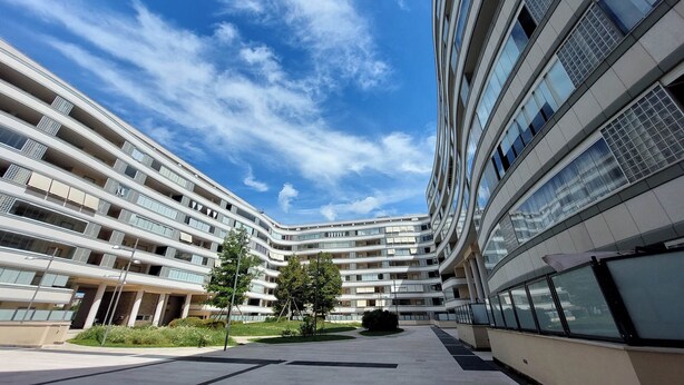 Um edifício moderno de vários andares com fachada curva, janelas amplas e um pátio central com árvores.