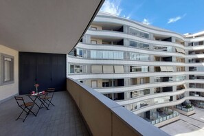 Ein mehrstöckiges Gebäude mit Balkonen, einer Terrasse mit Tisch und Stühlen und blauem Himmel.