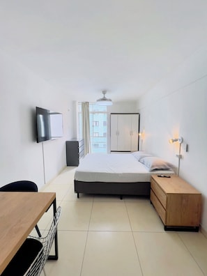 A hotel room with a bed, bedside table, lamp, TV, and a view of the outside. - Edifício Tiatã (São Paulo)